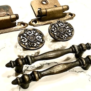 May include: Two antique brass-finished metal hinges, two round cabinet knobs with an intricate openwork scroll pattern, and two matching metal drawer pulls with a tapered, ribbed design and a weathered, dark metallic finish.