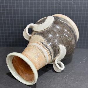 May include: A handmade ceramic vase with a wide base and a narrow neck. The vase features a speckled brown and tan glaze with white, curved handles. The interior of the neck is a warm orange color, and the rim is white.