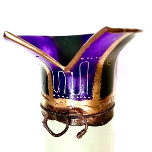 May include: A decorative, hand-painted vase with a flared top. The vase features a deep purple interior, outlined with gold and black accents. White, stylized patterns adorn the exterior, and a rope-like detail encircles the base.