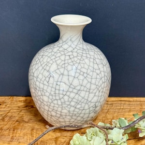 May include: A round ceramic vase with a narrow neck and a flared rim, featuring an off-white glaze with a prominent dark gray crackle pattern across the entire surface. The vase has a smooth, glossy finish and a bulbous, spherical body shape.