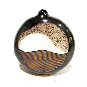 May include: A round, dark brown ceramic vase with a unique design. The vase features a white and speckled tan section, and a textured, brown, chevron-patterned lower portion. The vase has a small opening at the top.