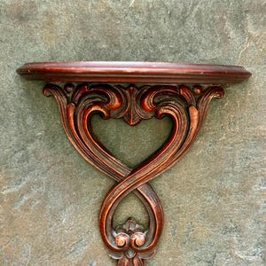 Puede incluir: Una estanter&iacute;a de pared de madera de color marr&oacute;n rojizo oscuro con detalles tallados ornamentados. La estanter&iacute;a tiene un borde frontal curvado y est&aacute; sostenida por un soporte decorativo con un dise&ntilde;o en forma de coraz&oacute;n. El soporte mide unos 30 cm de ancho.