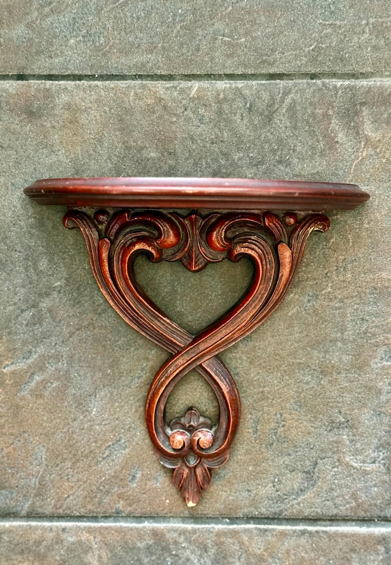 Puede incluir: Una estanter&iacute;a de pared de madera de color marr&oacute;n rojizo oscuro con detalles tallados ornamentados. La estanter&iacute;a tiene un borde frontal curvado y est&aacute; sostenida por un soporte decorativo con un dise&ntilde;o en forma de coraz&oacute;n. El soporte mide unos 30 cm de ancho.
