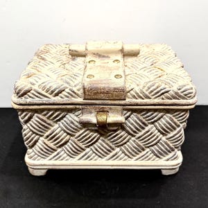 May include: A rectangular storage box with a textured, woven rope pattern in off-white and dark brown. The lid features a central latch with four small brass-colored studs. The box has a distressed finish and rests on four small, sturdy feet.