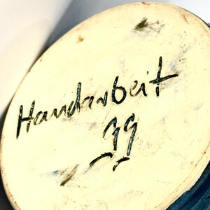 May include: The circular base of a ceramic object features a cream-colored surface with the handwritten black text Handarbeit and the number 99 inscribed in the center. The edges of the base show a dark blue glaze finish on the outer rim.