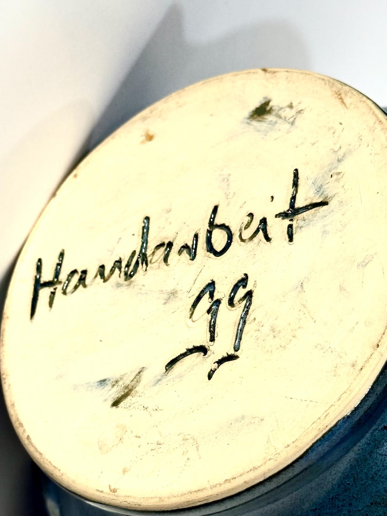 May include: The circular base of a ceramic object features a cream-colored surface with the handwritten black text Handarbeit and the number 99 inscribed in the center. The edges of the base show a dark blue glaze finish on the outer rim.