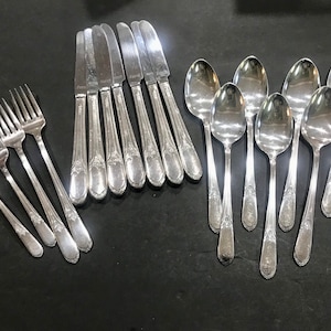 May include: A collection of silver-colored flatware, including forks, knives, and spoons, arranged on a dark surface. The silverware has decorative detailing on the handles. The spoons have a rounded bowl shape.