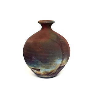 May include: A small, round ceramic vase with a narrow neck and a flared rim. The surface features a mottled, textured finish with blended tones of deep reddish-brown, earthy charcoal, and patches of muted blue and cream near the base.