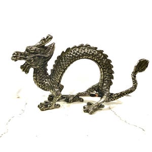 May include: A silver-toned metal dragon figurine featuring a textured, scaled body, a curved arched back, and a detailed head with open jaws. The creature is depicted in a standing position with four clawed feet and a long, slender tail.