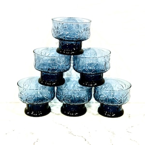 May include: A stack of seven blue glass dessert bowls. Each bowl has a round, floral-patterned top and a dark blue pedestal base. The bowls are arranged in a pyramid shape, with one bowl at the top and six bowls below.