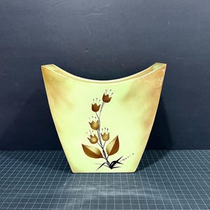 May include: A pale yellow ceramic vase with a wide, concave top edge and a tapered base. The front surface features a central graphic illustration of a stylized plant with brown, bell-shaped flowers and pointed leaves extending upward.