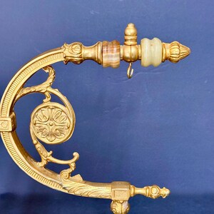 May include: A gold-toned metal bracket featuring an ornate curved arm with a circular floral medallion. The horizontal extension includes two smooth, banded stone accents in shades of brown and cream, with a small metal hook hanging underneath.