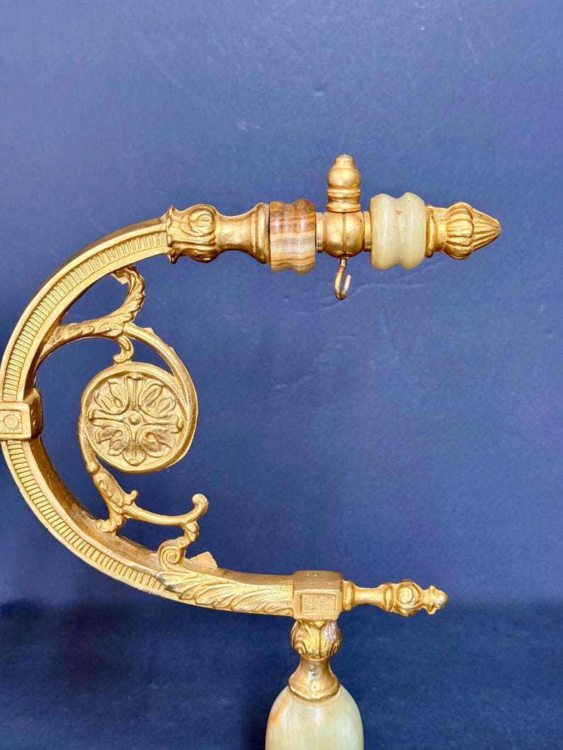 May include: A gold-toned metal bracket featuring an ornate curved arm with a circular floral medallion. The horizontal extension includes two smooth, banded stone accents in shades of brown and cream, with a small metal hook hanging underneath.