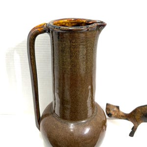 May include: A brown ceramic pitcher with a handle and a spout. The pitcher has a bulbous base and a tall, cylindrical body. The handle is a darker brown color. A small, bronze-colored decorative object is to the right of the pitcher.