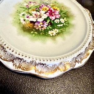 May include: A round porcelain plate featuring a central floral bouquet with pink, white, and yellow flowers against a green background. The plate has a scalloped edge with a raised beaded texture and an ornate gold filigree trim along the border.