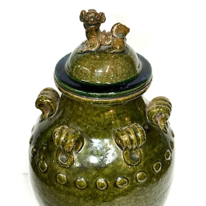 May include: A dark olive green glazed ceramic jar with a matching lid topped by a small sculpted animal figure. The jar features a series of raised circular dots around the shoulder and four decorative scroll-shaped handles on the upper body.