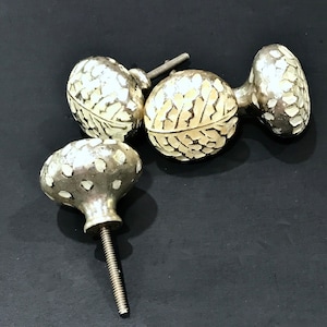 May include: Four decorative cabinet knobs with a silver finish and a textured, off-white inlay design. The knobs have a variety of shapes, including round and oval, and are set against a dark background.
