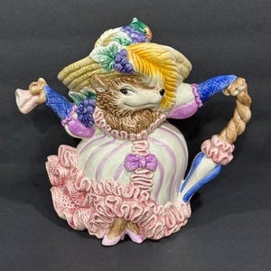 May include: A whimsical ceramic teapot featuring a hedgehog character dressed in a Victorian-style outfit. The teapot is primarily white with pink and purple accents, a straw hat, and a decorative handle. The teapot is approximately 10 inches tall.