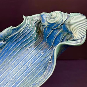 May include: A ceramic bowl with a deep blue and light green glaze, featuring a textured surface with raised vertical lines and decorative shell-like formations along the rim. The piece has an organic, wavy edge and a glossy, mottled finish.