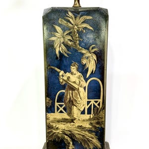 May include: A rectangular table lamp with a blue background and a gold-colored illustration of a person playing a stringed instrument under palm trees. The lamp has a brass-colored base and a light fixture.