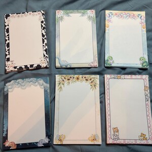 May include: Six rectangular memo pads featuring various decorative borders. Designs include a black and white cow print, a tropical leaf frame with dinosaurs, a pastel bubble border with turtles, a starry night sky, a floral wreath, and a pink striped frame.