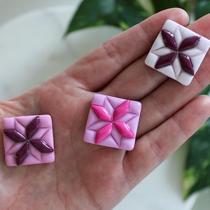 May include: Three square buttons featuring a raised, four-pointed star pattern in the center. One button is white with dark plum star accents, while the other two are shades of pink with either dark plum or bright magenta star details.