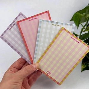 May include: Four rectangular notepad sheets featuring a gingham check pattern in purple, pink, light blue, and yellow. Each sheet has a border and the text small notes printed in the top corner, with a small fish logo on the yellow sheet.