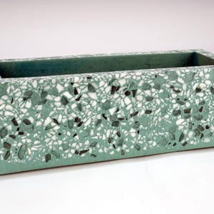 Könnte beinhalten: Ein rechteckiger Pflanzkübel mit einer salbeigrünen Basis und einem gesprenkelten Terrazzo-Muster aus weißen und dunkelgrauen Steinfragmenten. Der Behälter hat einen glatten, flachen oberen Rand und einen hohlen Innenraum.