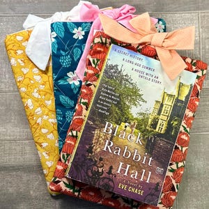 May include: A stack of colorful book covers with matching bows. The covers feature floral and patterned designs in yellow, teal, and red. The book "Black Rabbit Hall" by Eve Chase is visible, with a cover featuring a house and garden.