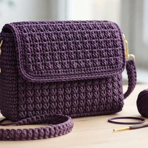 May include: A deep purple crochet crossbody bag featuring a textured stitch pattern, a rectangular flap, and a matching crochet shoulder strap attached with gold-tone metal clasps. The bag is accompanied by a ball of purple yarn and a crochet hook.