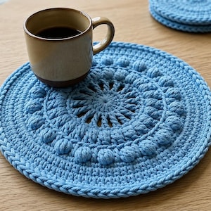 May include: A light blue circular crochet placemat featuring a textured bobble stitch pattern and a central radiating design. A ceramic mug with dark liquid sits on the placemat, and a matching smaller circular crochet coaster is nearby.
