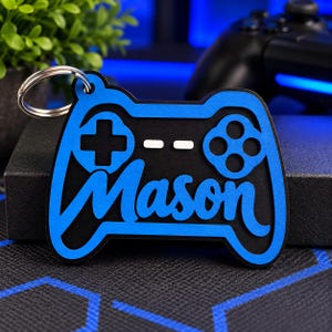 May include: A blue and black layered keychain shaped like a video game controller. The design features a cutout D-pad, action buttons, and two horizontal bars. The name Mason is written in a blue script font across the center of the controller.