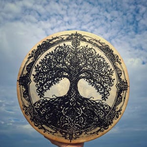 May include: A circular drum featuring a detailed black ink illustration of a tree with intricate, sprawling roots and branches. The design is enclosed within a decorative border, set against a light tan surface with a natural texture.