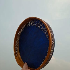May include: A circular tambourine featuring a deep blue drumhead surface and a wooden frame. The outer edge of the frame is decorated with a dark band containing white circular patterns. Metal rings are attached along the inner rim.