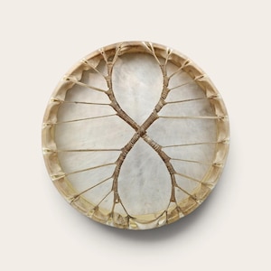 Pode incluir: Um tambor de moldura circular com uma pele animal translúcida de cor branco-sujo esticada sobre um aro de madeira. A pele do tambor é fixada por uma rede complexa de finos cordões de couro castanho claro que formam um padrão em X.