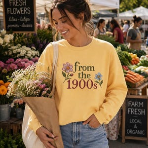May include: A yellow long-sleeve crewneck sweatshirt featuring a cross-stitch style graphic of a pink flower, a blue flower, and the dark green and maroon text "from the 1900s" centered on the chest. The sweatshirt is paired with light blue denim jeans.