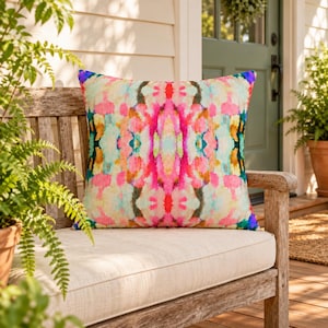 Könnte beinhalten: Ein quadratisches Zierkissen mit einem lebendigen, symmetrischen abstrakten Muster aus vermischten Farbspritzern in Pink, leuchtendem Gelb, Türkis und Creme. Das Design zeigt einen gespiegelten Aquarell-Effekt auf der Stoffoberfläche.