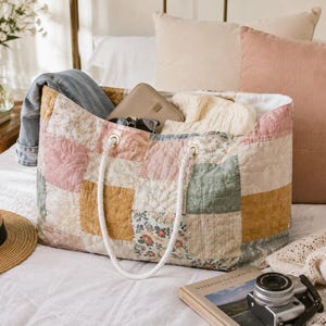 May include: A large rectangular quilted tote bag featuring a patchwork design of muted pink, mustard yellow, sage green, and floral fabric squares. The bag has two white braided rope handles attached with gold-toned metal grommets.