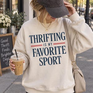 Könnte beinhalten: Ein cremefarbenes Sweatshirt mit Rundhalsausschnitt und dem dunkelblauen Schriftzug "THRIFTING IS MY FAVORITE SPORT", wobei das Wort "IS" von zwei horizontalen roten Linien eingerahmt wird. Das Kleidungsstück hat lange Ärmel.
