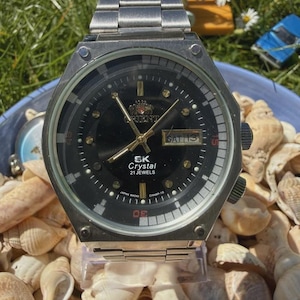 Könnte beinhalten: Eine silberfarbene Orient SK Crystal Armbanduhr mit schwarzem Zifferblatt, goldfarbenen Zeigern und einem Edelstahl-Gliederarmband. Das Zifferblatt zeigt das Orient-Logo, den Schriftzug SK Crystal 21 Jewels und eine Datumsanzeige mit SAT 13.