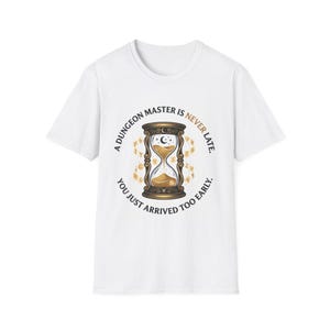 Puede incluir: Una camiseta blanca de manga corta con un gráfico central de un reloj de arena con arena dorada. El texto dice: A DUNGEON MASTER IS NEVER LATE. YOU JUST ARRIVED TOO EARLY. El diseño incluye pequeños símbolos dorados alrededor del reloj.