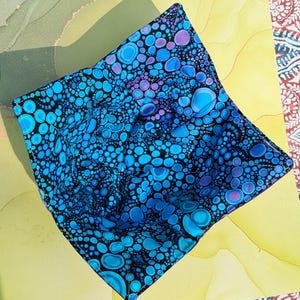 May include: A square fabric bowl cozy featuring a black background with a vibrant pattern of circular shapes in shades of bright blue, cyan, and purple. The quilted textile accessory has stitched seams forming a structured, four-cornered shape.