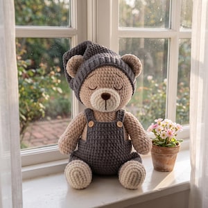 May include: A crocheted teddy bear with light tan yarn, wearing a dark gray ribbed nightcap and matching dark gray overalls with two small wooden buttons. The bear has closed eyes, a dark brown stitched nose, and a light tan muzzle.