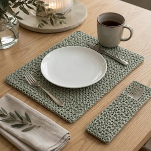 Op de afbeelding: Een saliegroene gehaakte rechthoekige placemat en een bijpassende rechthoekige bestekhouder. De set bevat een wit keramieken bord, een zilveren vork, een zilveren mes en een mok gevuld met een donkere drank op een houten ondergrond.
