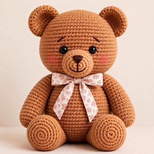 Puede incluir: Un oso de peluche tejido a ganchillo de color marrón con ojos de seguridad negros, nariz bordada en marrón oscuro y mejillas sonrosadas. El oso lleva un lazo de color claro atado al cuello, decorado con un pequeño estampado floral rosa y verde.
