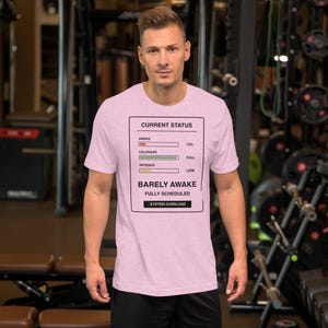 Puede incluir: Una camiseta de manga corta de color rosa claro con un recuadro gráfico negro que contiene el texto CURRENT STATUS, AWAKE 12%, CALENDAR FULL, PATIENCE LOW, BARELY AWAKE, FULLY SCHEDULED y SYSTEM OVERLOAD junto a barras de progreso.