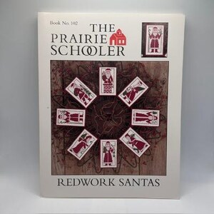 Può includere: Un libro di modelli con copertina flessibile intitolato The Prairie Schooler Book No. 102 Redwork Santas. La copertina presenta un'immagine centrale di otto piccoli disegni rettangolari a punto croce di Babbo Natale in filo rosso.