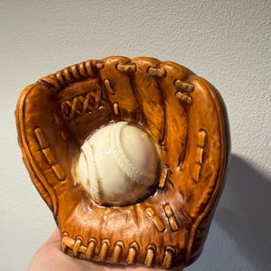 Peut inclure: Une sculpture en céramique représentant un gant de baseball avec une finition marron brillant et une balle de baseball couleur crème au centre. Le gant présente des détails moulés imitant les coutures, les lacets et les doigts du cuir.