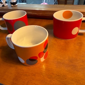 May include: Three ceramic mugs with white interiors and handles. One mug is red with large grey circles, one is orange with overlapping red, grey, and white circles, and one is red with a large orange circle inside and a white circle on the exterior.