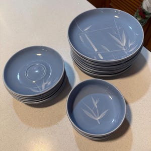 May include: A collection of light blue ceramic dinnerware including a stack of large dinner plates, a stack of saucers, and a stack of small bowls. Each piece features a white painted bamboo leaf design on the surface of the smooth, round dishes.
